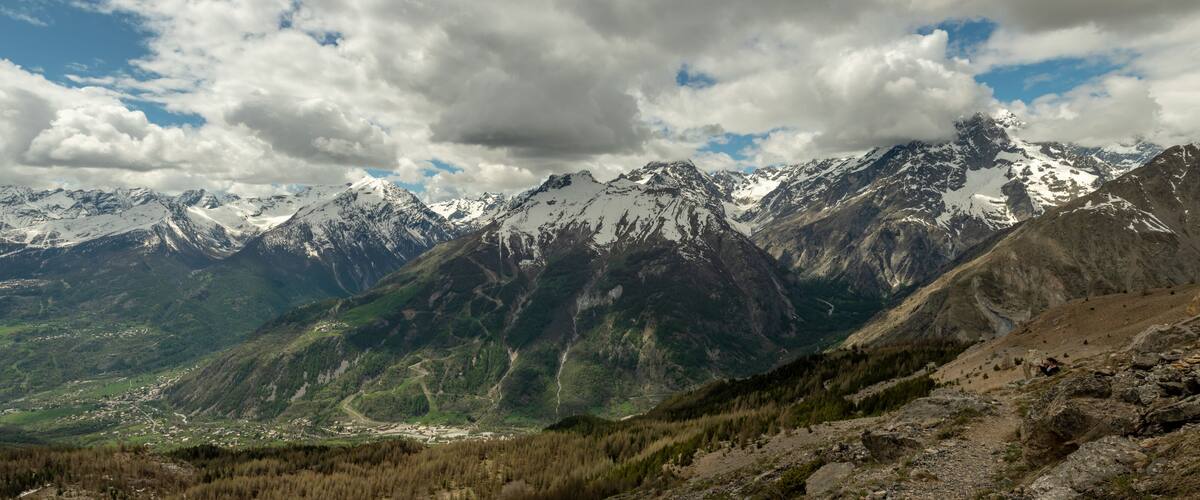 A mountainous landscape with snow-capped peaks, a valley with a small settlement, and a clear sky with clouds.