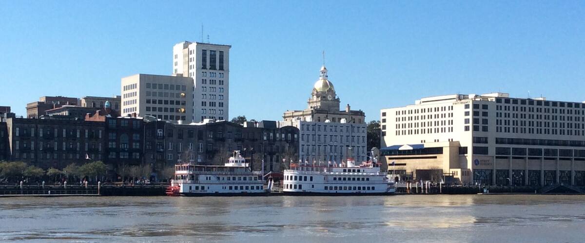 A river with two boats, a city skyline with historic and modern buildings, and a clear blue sky.