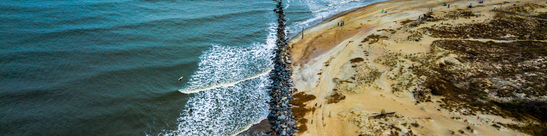 Vista aérea de uma praia com um molhe rochoso, ondas e uma orla arenosa.