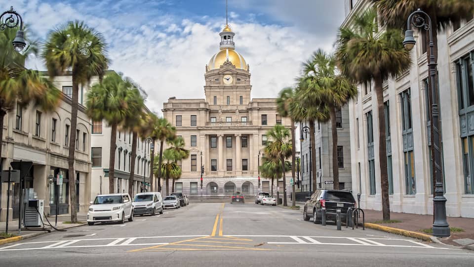 A street with parked cars, palm trees, and a large building with a golden dome.