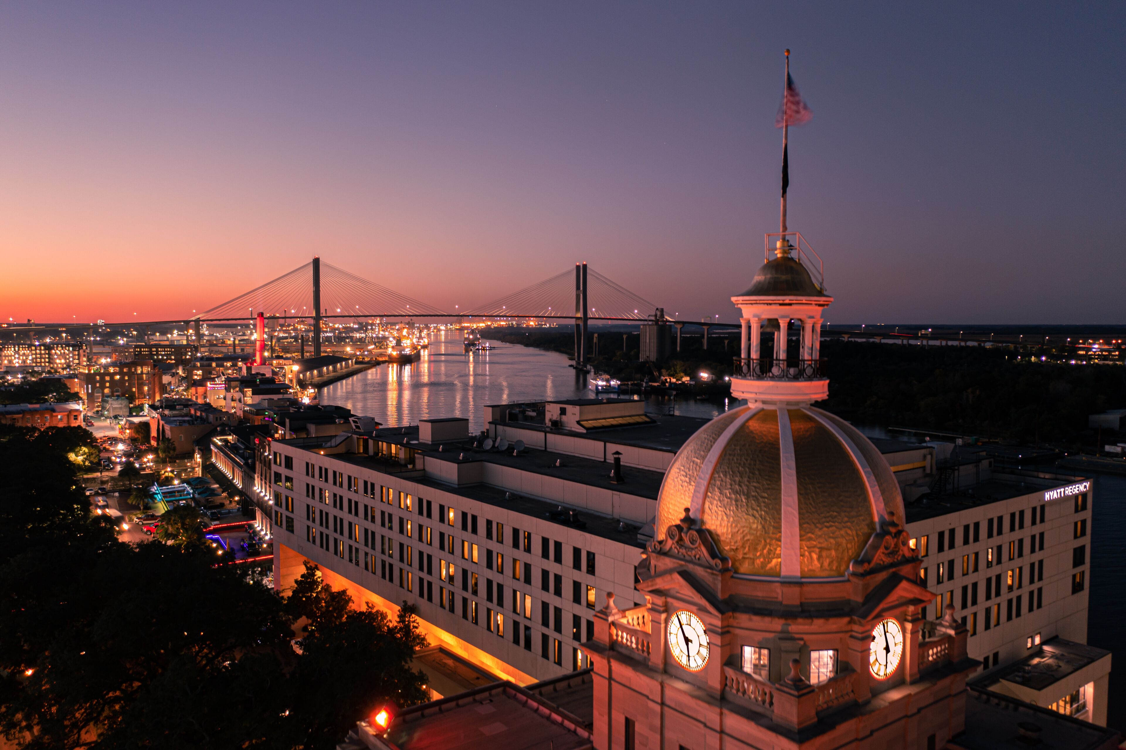 A cityscape at dusk with a prominent building featuring a clock tower and a dome, a bridge in the background, and a river.