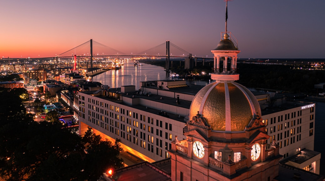A cityscape at dusk with a prominent building featuring a clock tower and a dome, a bridge in the background, and a river.