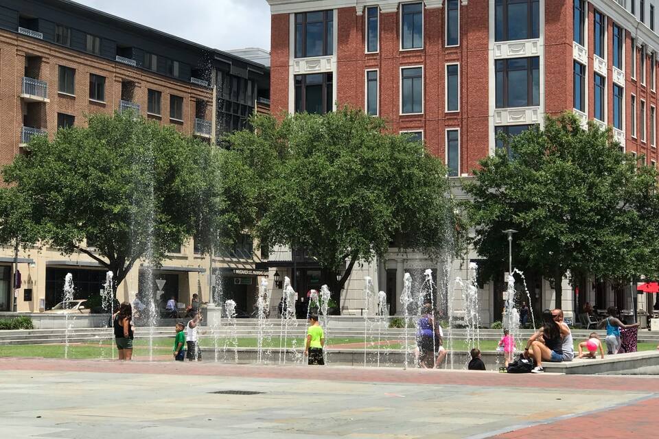 Ein öffentlicher Platz mit einem Springbrunnen, Bäumen und Gebäuden im Hintergrund.