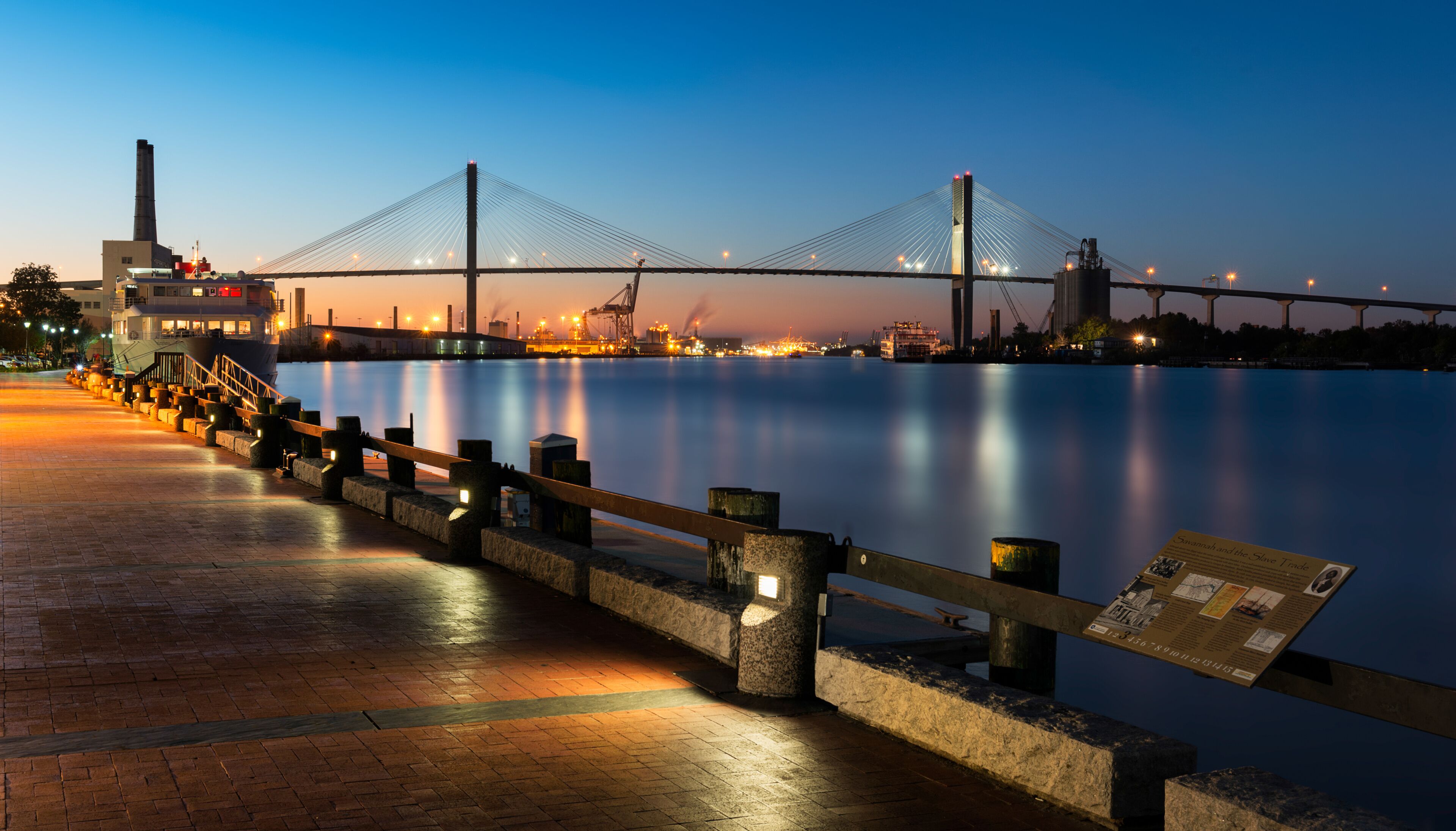A bridge at dusk with a calm river, a dock with a boat, and an industrial area in the background.