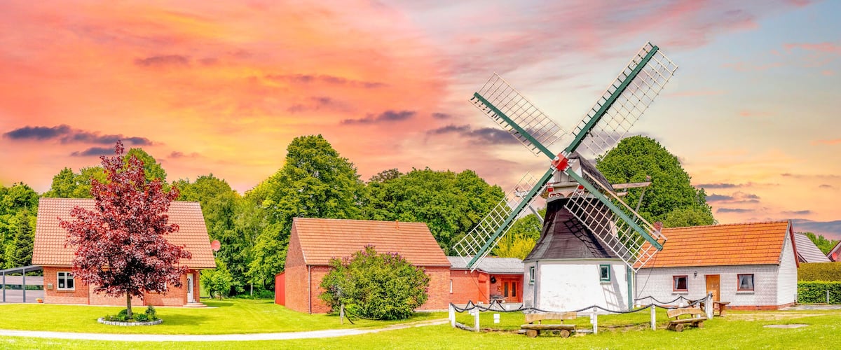 A traditional windmill in a rural setting with red-roofed buildings and lush greenery.