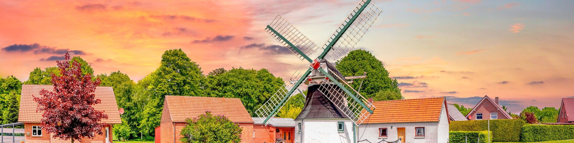 A traditional windmill in a rural setting with red-roofed buildings and lush greenery.