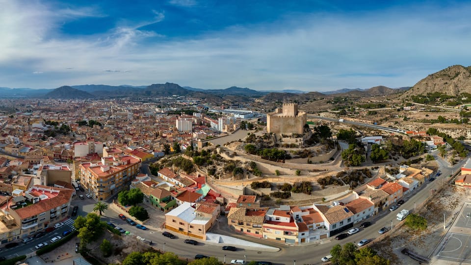 A panoramic view of a city with a prominent castle on a hill, surrounded by mountains and a mix of urban and rural landscapes.