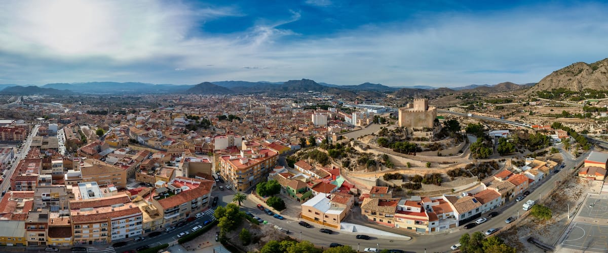 A panoramic view of a city with a prominent castle on a hill, surrounded by mountains and a mix of urban and rural landscapes.