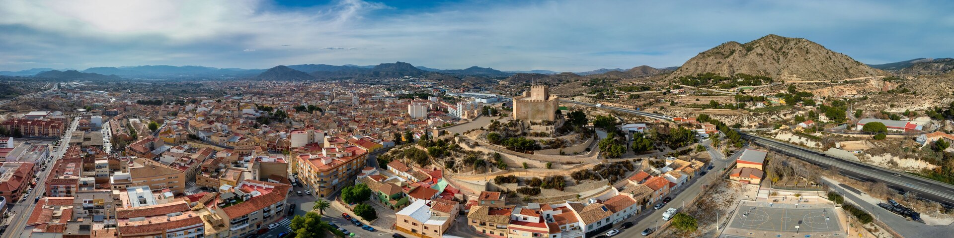 A panoramic view of a city with a prominent castle on a hill, surrounded by mountains and a mix of urban and rural landscapes.