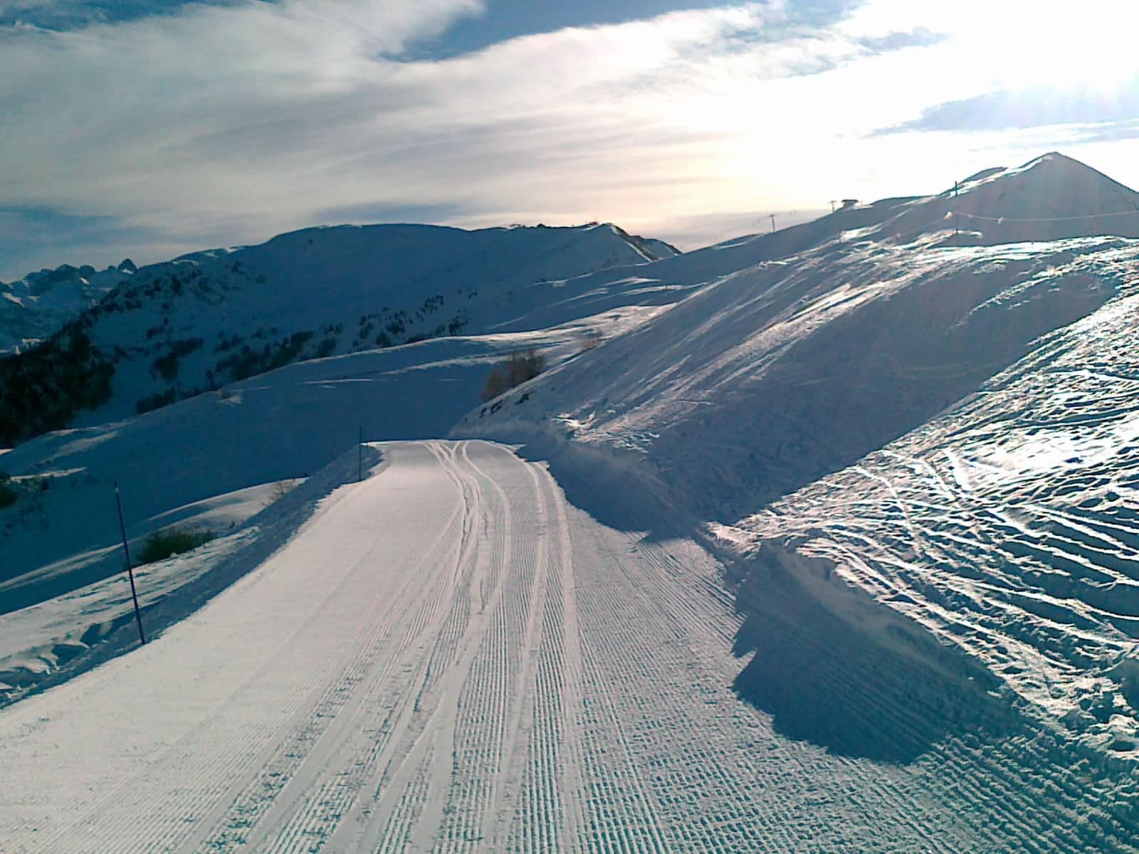 Estrada montanhosa coberta por neve, com marcas de pneus, cercada por encostas nevadas e céu claro.