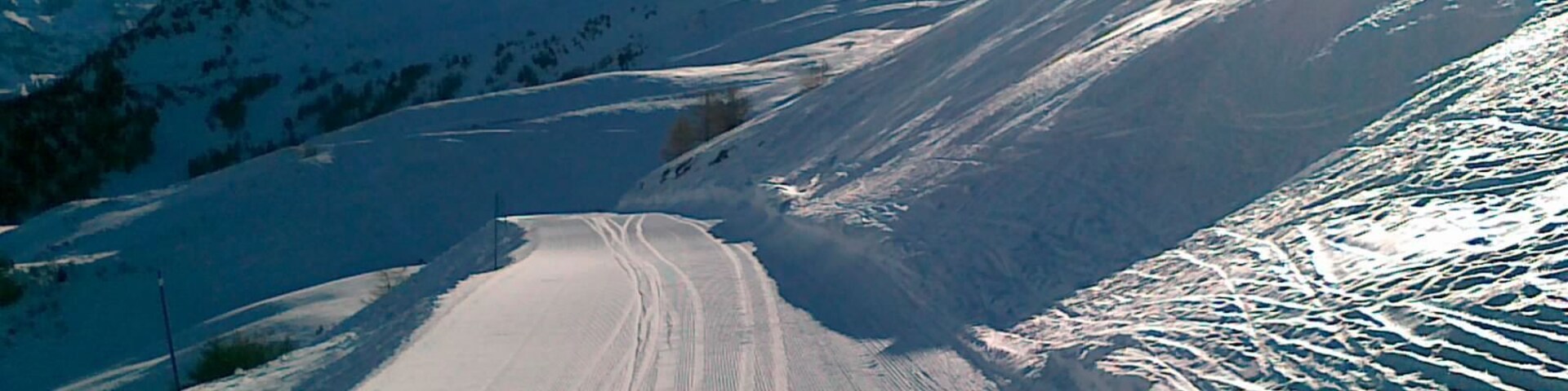 Estrada montanhosa coberta por neve, com marcas de pneus, cercada por encostas nevadas e céu claro.