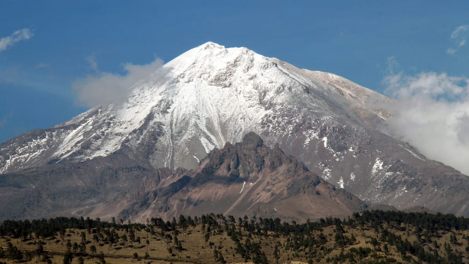 A snow-capped mountain with a clear sky and a forested valley below.