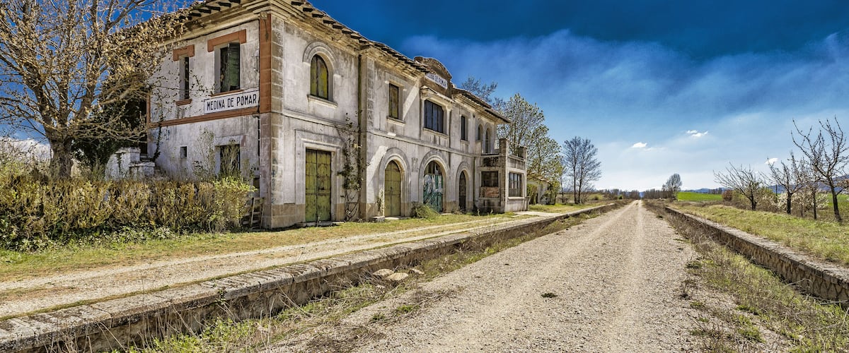 An old building named 'Medina de Pomar' with a dirt road leading to it, surrounded by trees and fields.