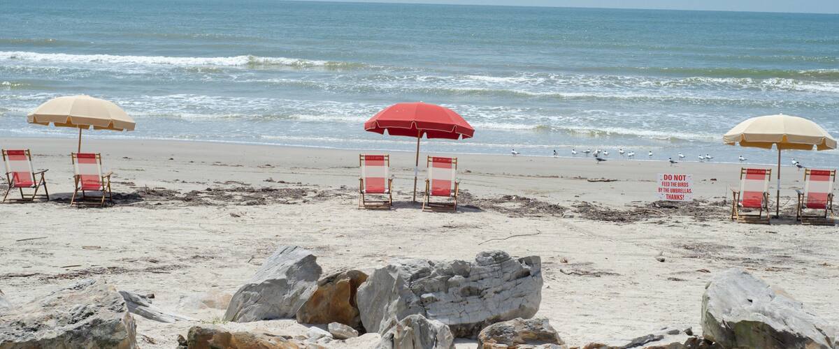 A beach with red and beige umbrellas, white and red beach chairs, and rocky outcrops.