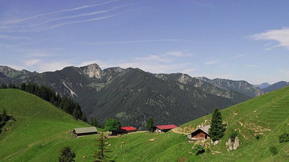 A mountainous landscape with green fields, a few buildings, and distant peaks.