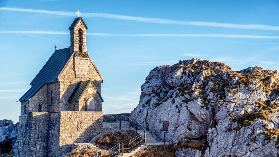 A stone church with a pointed roof and a bell tower, surrounded by rocky terrain and a clear sky.