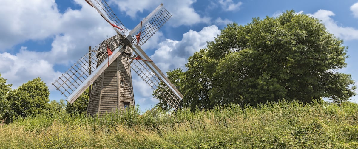 A traditional windmill with wooden structure and metal blades, surrounded by lush greenery and a clear blue sky.