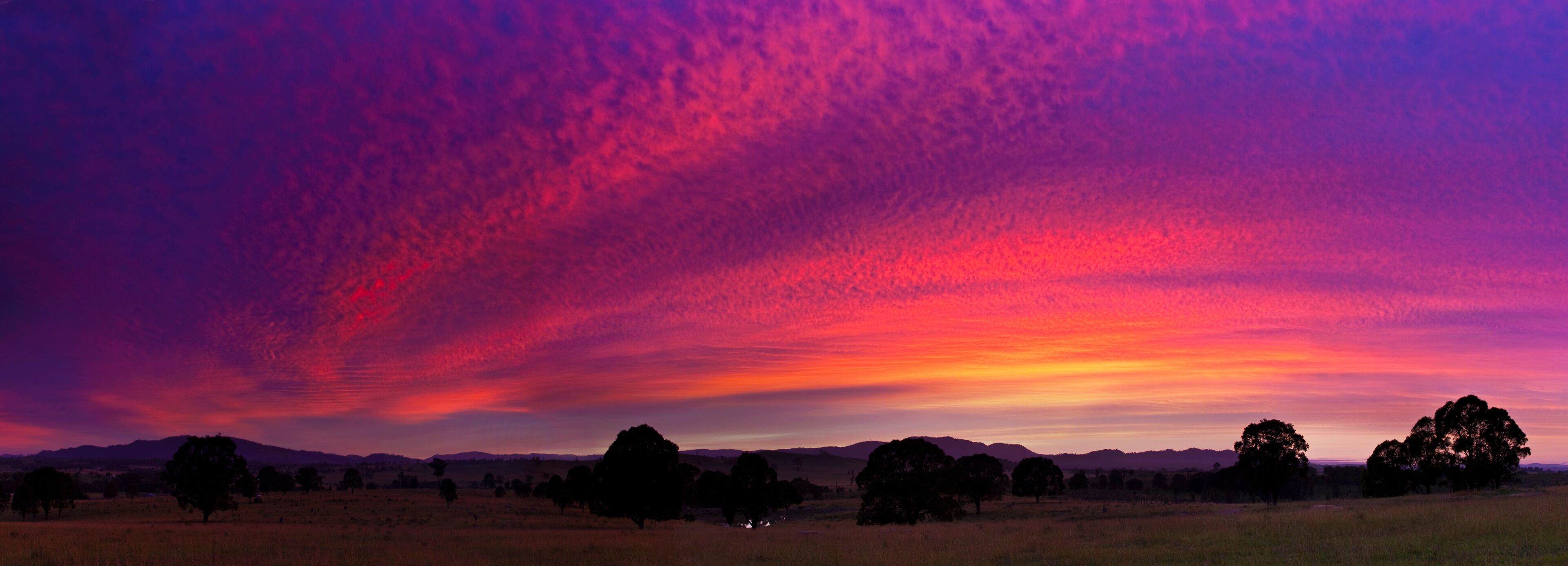 Um pôr do sol sobre um campo gramado com árvores e colinas ao longe.