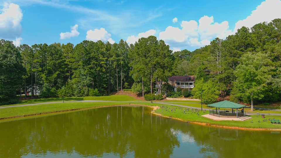 A serene lake surrounded by lush greenery, a gazebo, and a two-story house with a balcony.