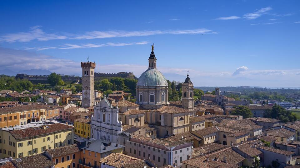 A historic cityscape with prominent domed and bell tower structures, surrounded by terracotta-roofed buildings.