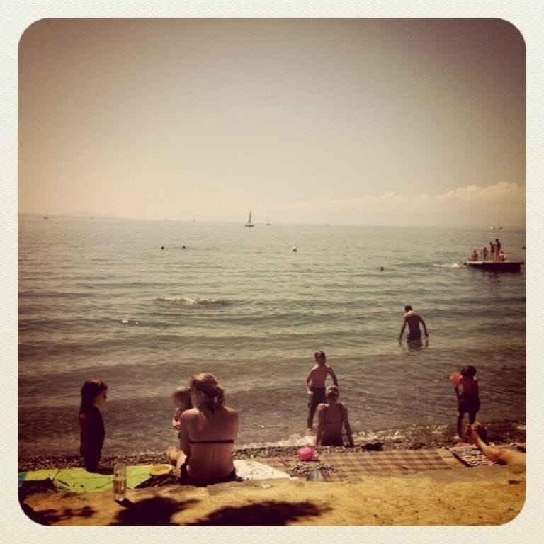 Una playa con gente sentada y de pie cerca del agua, un velero a lo lejos y un bote peque ño a la derecha.