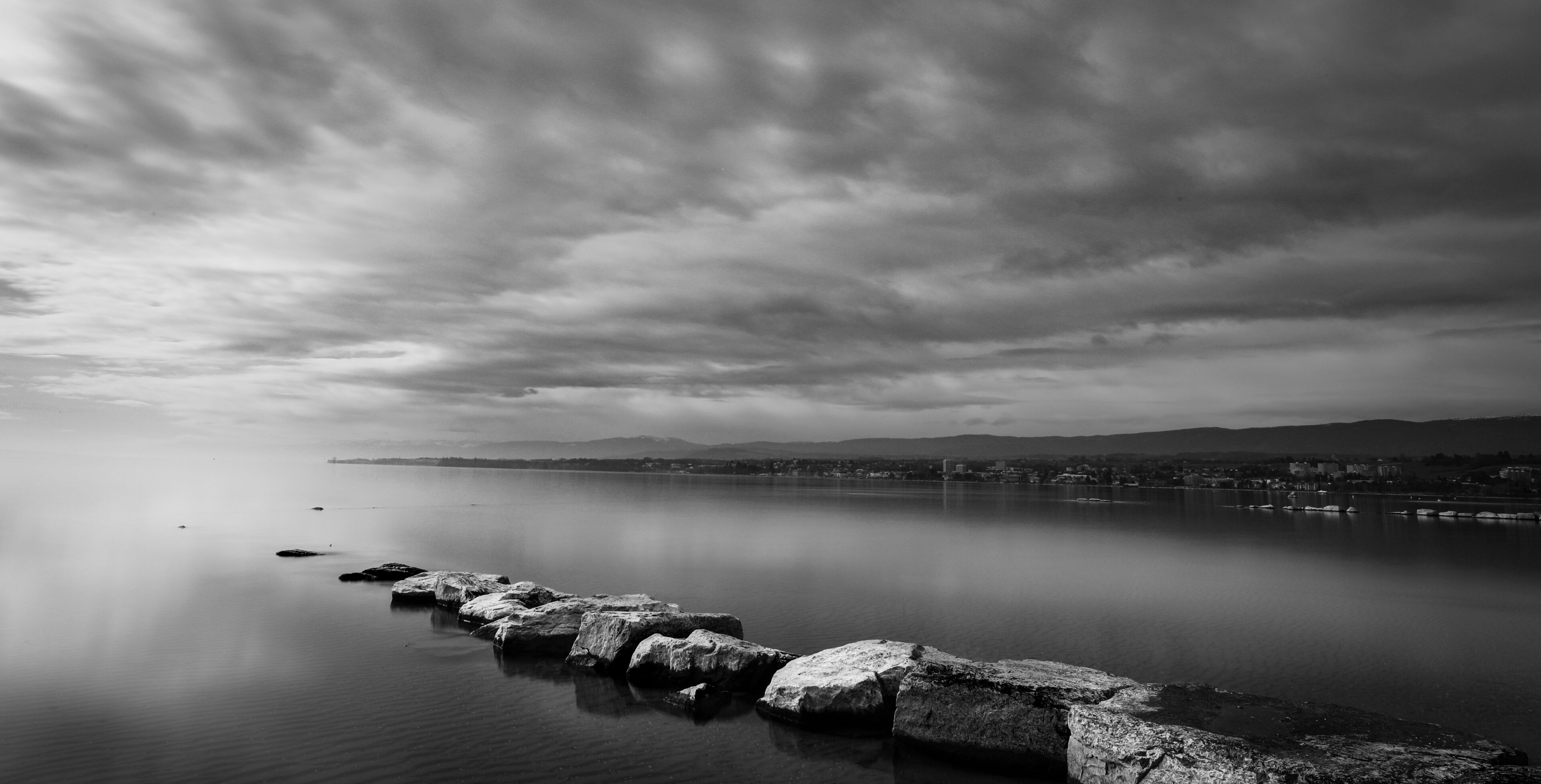 A calm lake with a row of rocks in the foreground and a cityscape in the background under a cloudy sky.