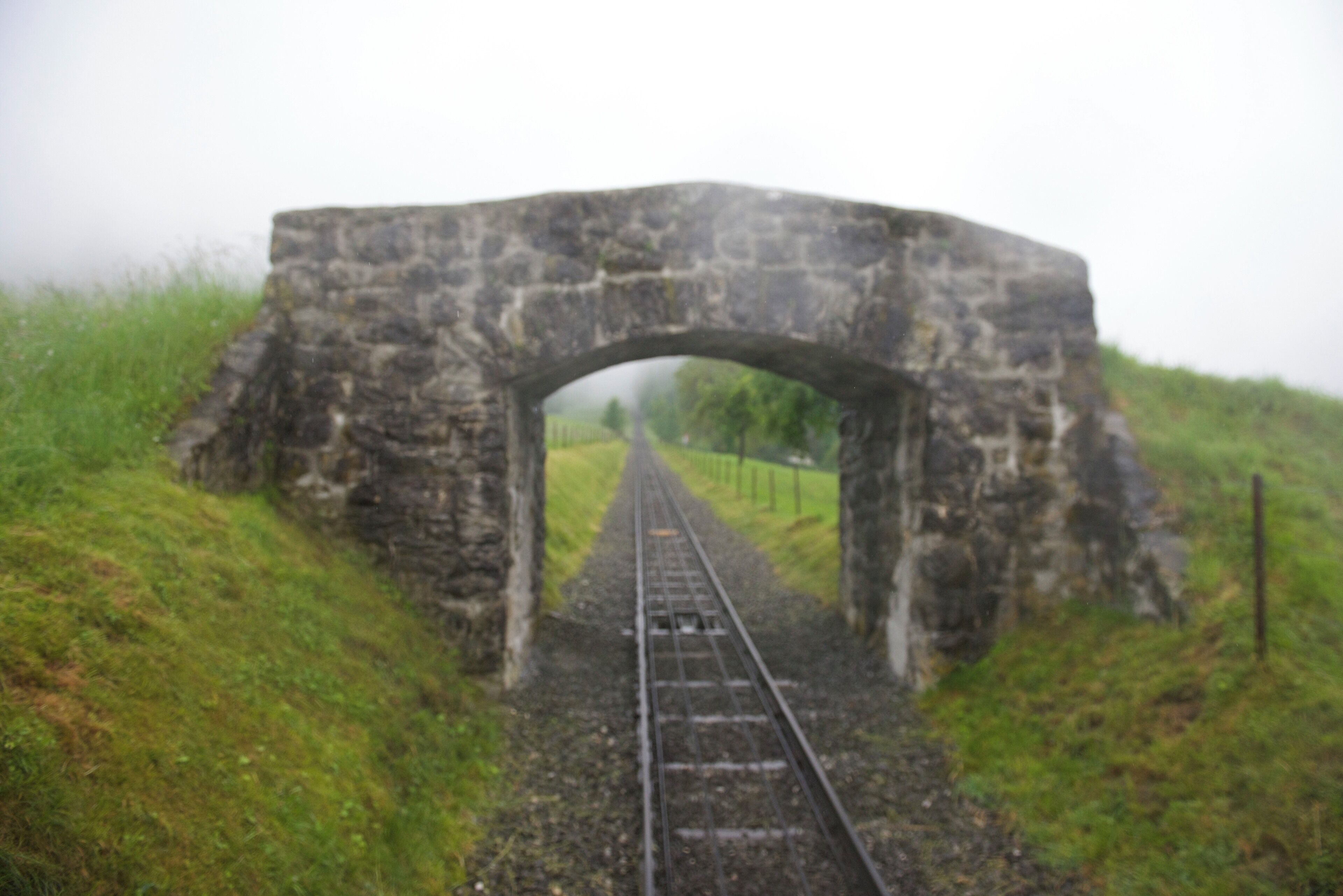 A stone railway bridge over tracks surrounded by grassy hills.
