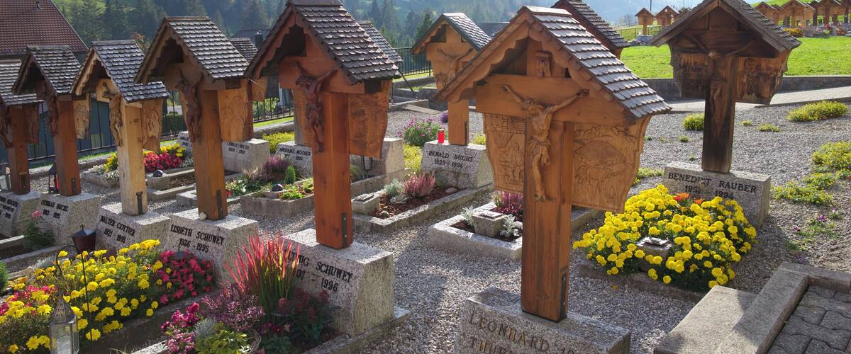 Wooden tombs with inscriptions, surrounded by colorful flowers and a mountainous backdrop.