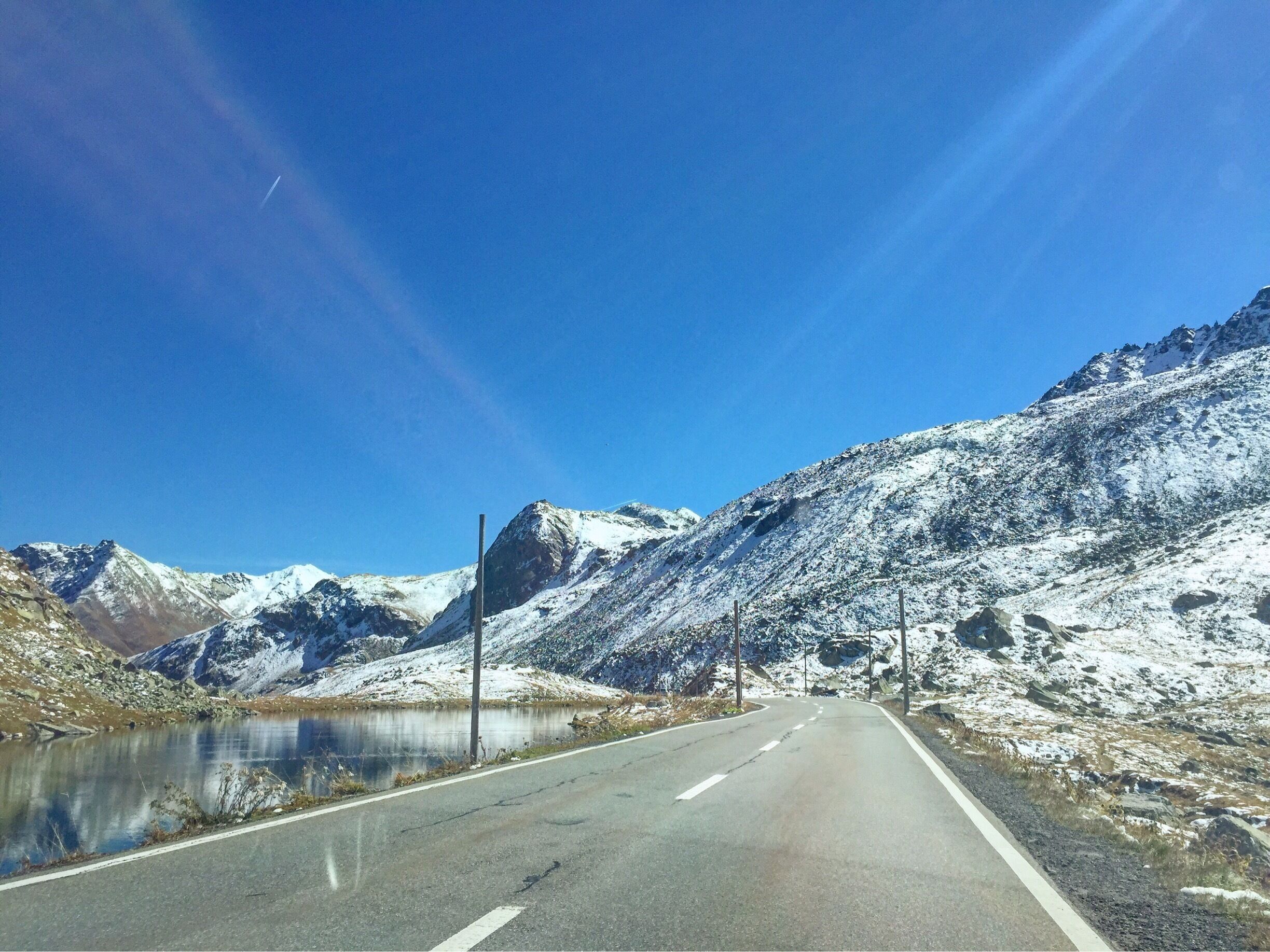 A snow-covered mountain road with a clear blue sky and a lake on the left.