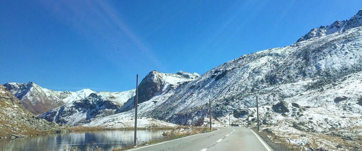 A snow-covered mountain road with a clear blue sky and a lake on the left.