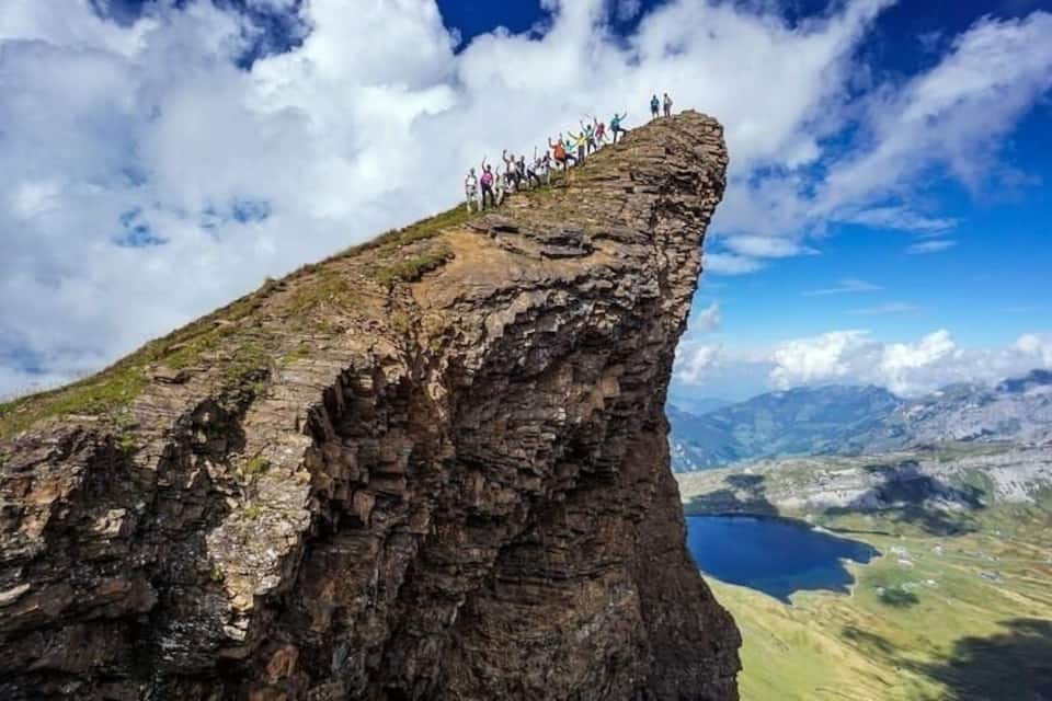 Un grupo de personas al borde de un acantilado rocoso, con vista a un valle y un lago.