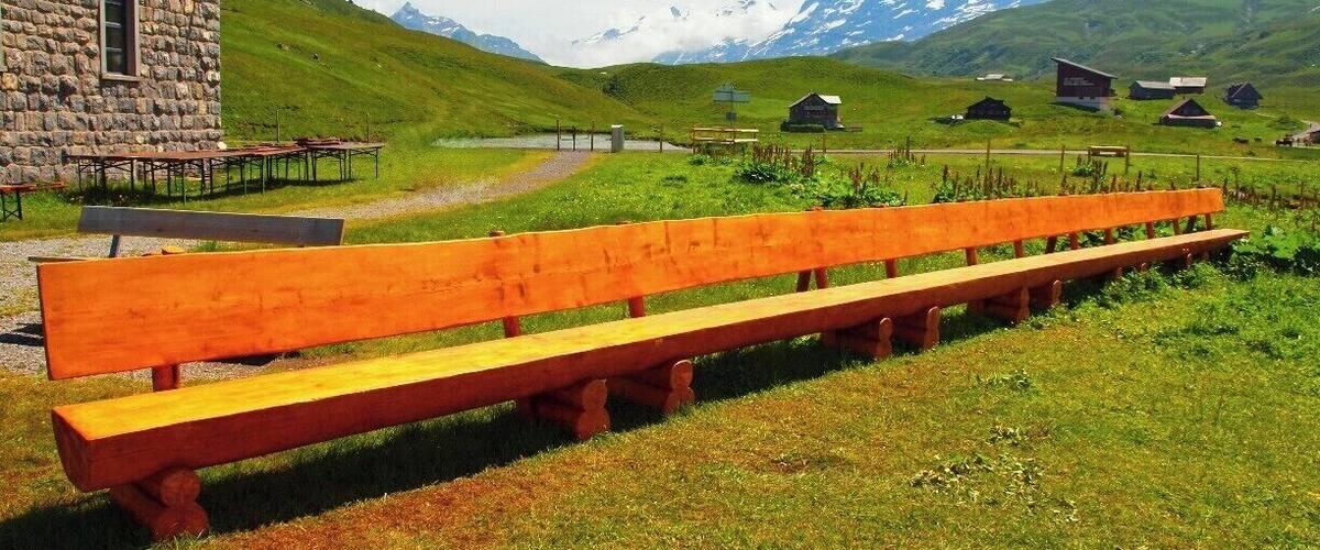 A wooden bench in a grassy field with a stone building and houses in the background, overlooking a mountain range.
