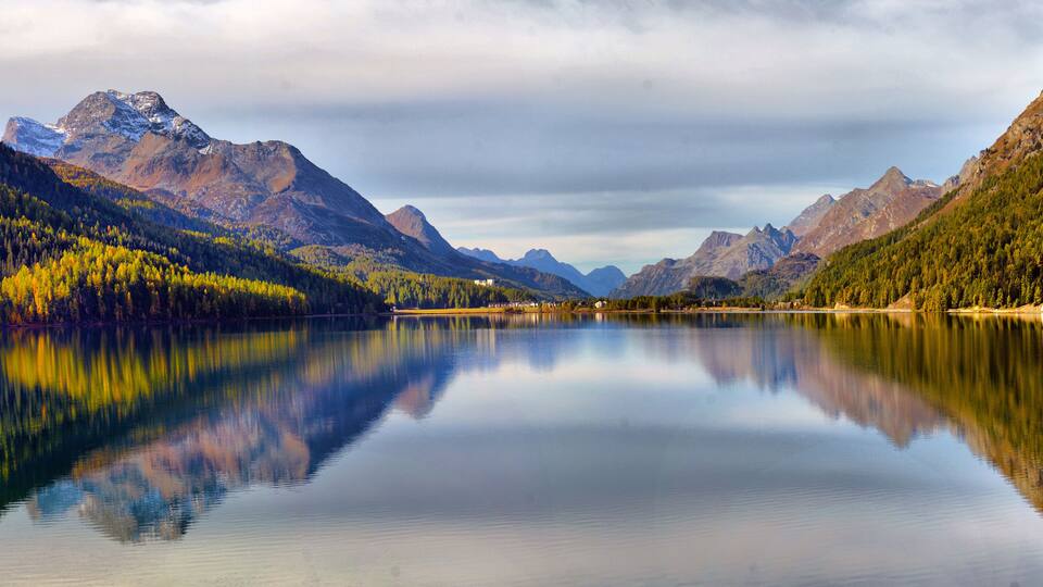 A calm lake surrounded by mountains with autumn foliage, reflecting the sky and landscape.