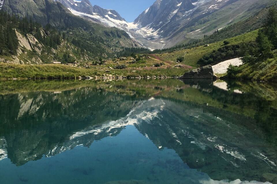 A clear mountain lake with reflections of the surrounding peaks and a small house near the water's edge.