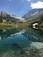 A clear mountain lake with reflections of the surrounding peaks and a small house near the water's edge.