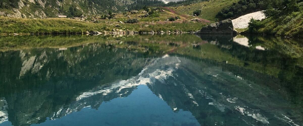 A clear mountain lake with reflections of the surrounding peaks and a small house near the water's edge.