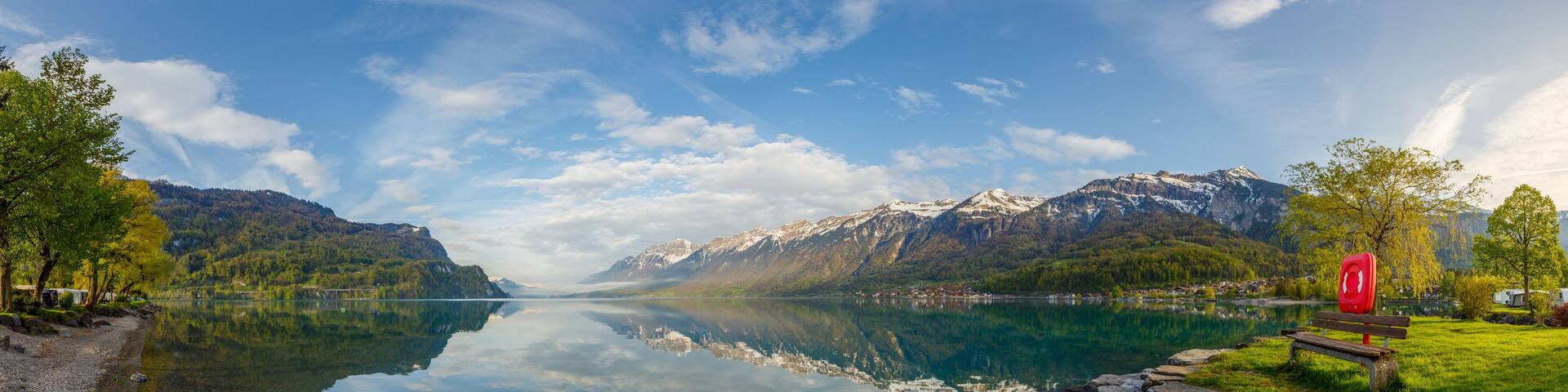 A calm lake surrounded by mountains, a clear sky, and a red lifebuoy on a bench.