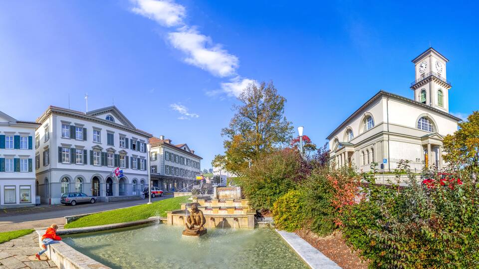 A historic town square with a fountain, classical architecture, and a clock tower.