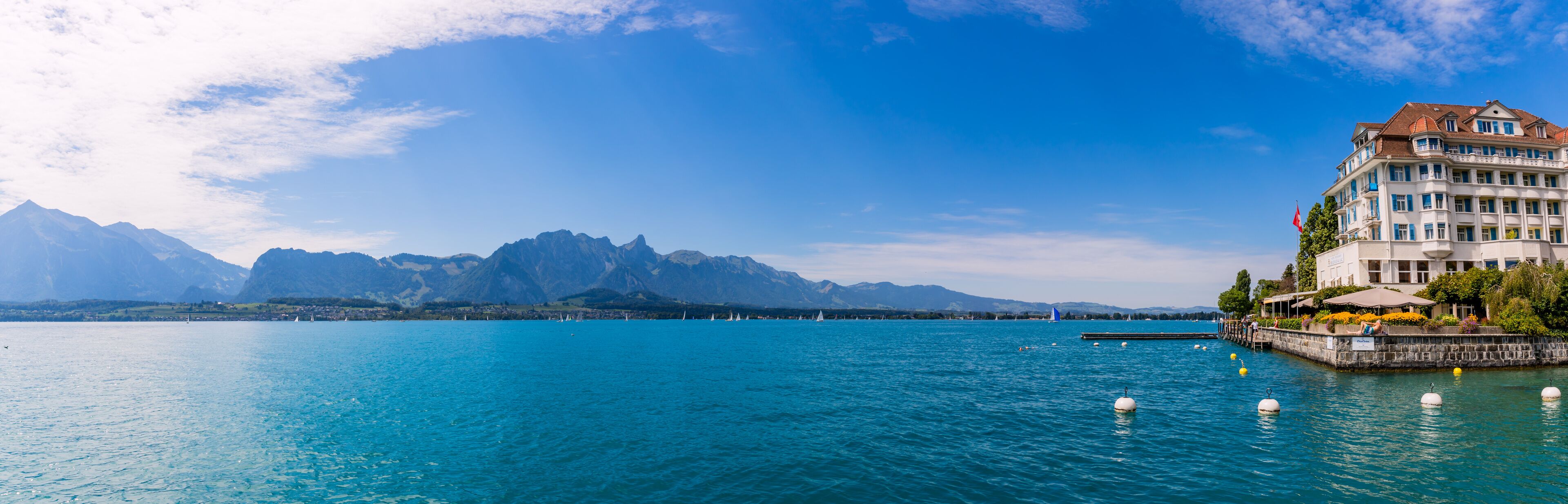 A lake with sailboats, a large building with a red flag, and mountains in the background.
