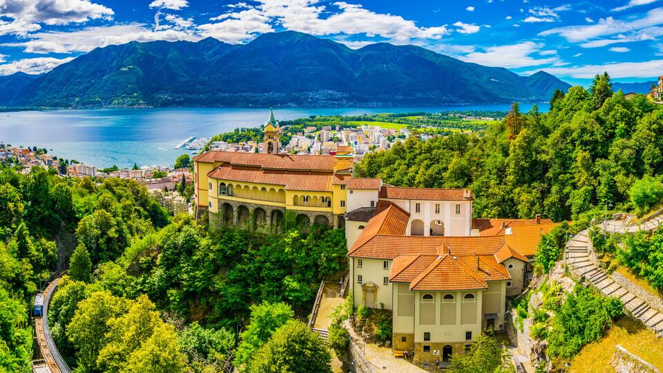 A scenic view of a town with a prominent yellow building with arches, surrounded by lush green hills and a lake with mountains in the background.