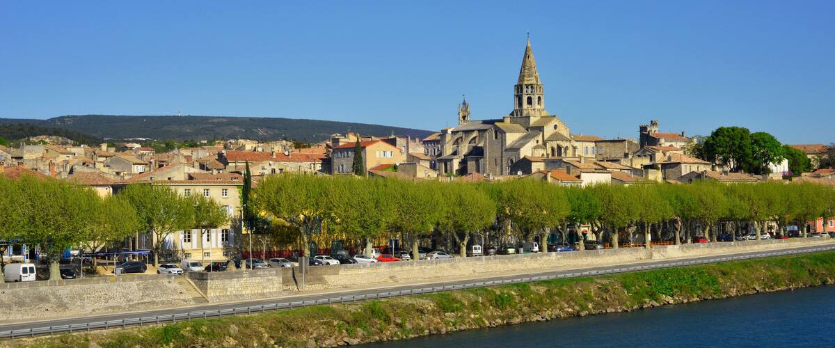A riverside town with historic buildings, a prominent church with a tall spire, and a row of trees along the waterfront.