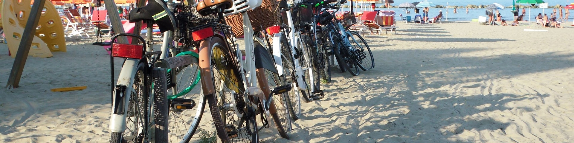 Bicycles parked on a sandy beach with umbrellas and beach chairs in the background.