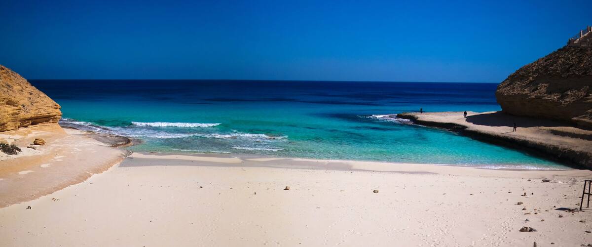A coastal landscape with a sandy beach, clear turquoise waters, and rocky cliffs.