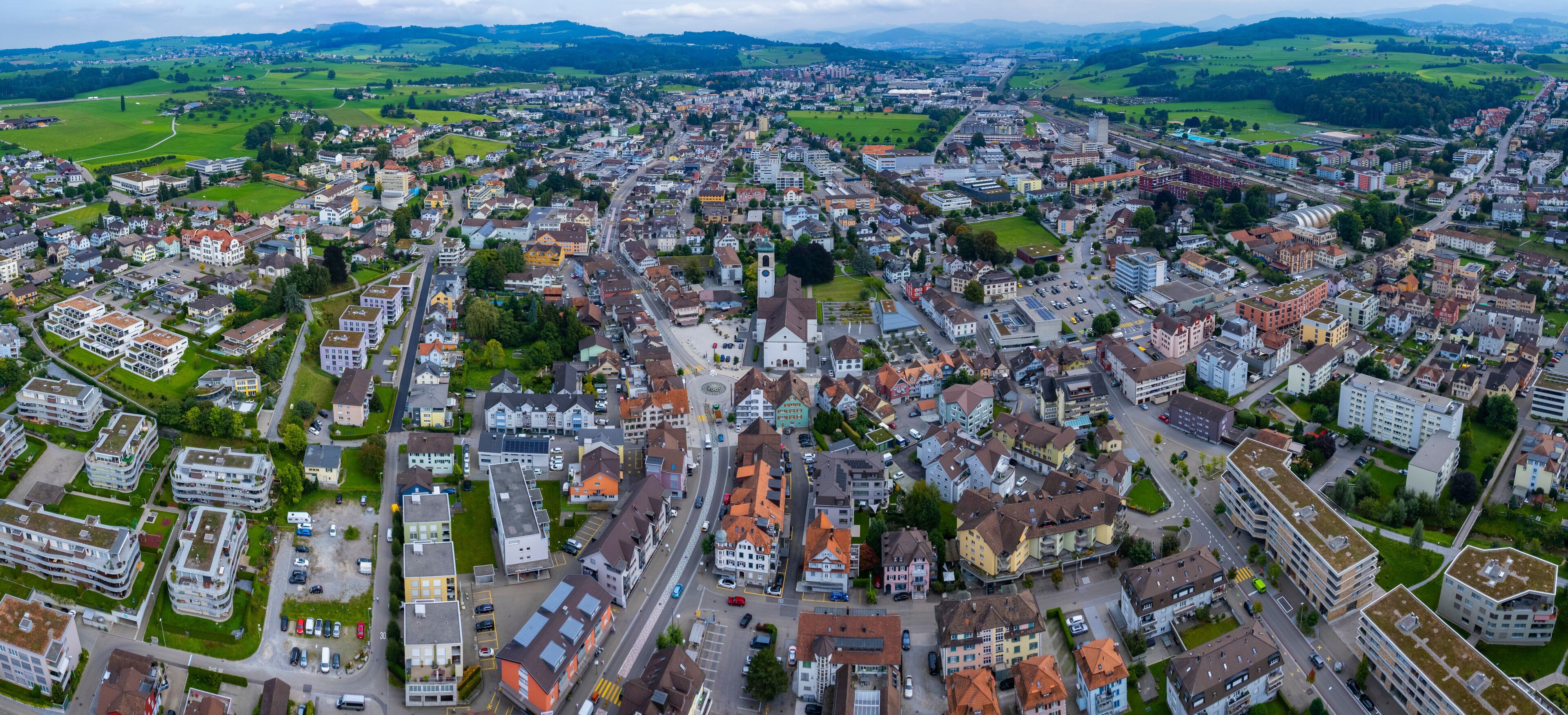 An aerial view of a town with a mix of residential and commercial buildings, a prominent church, and surrounding green fields.