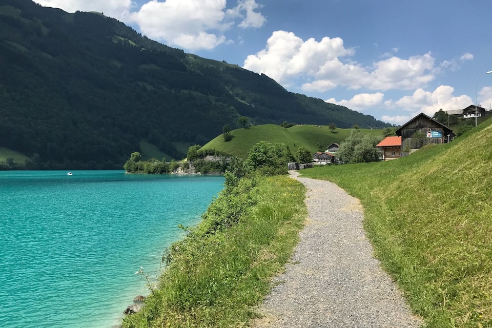 A gravel path beside a turquoise lake, surrounded by green hills and a wooden house with a red roof.