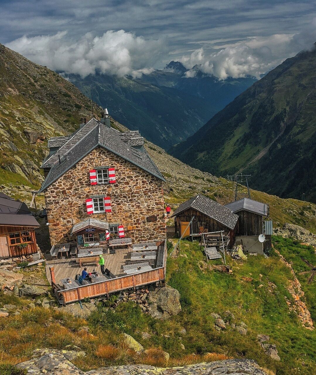 A stone house with red shutters, wooden structures, and a wooden deck in a mountainous area.