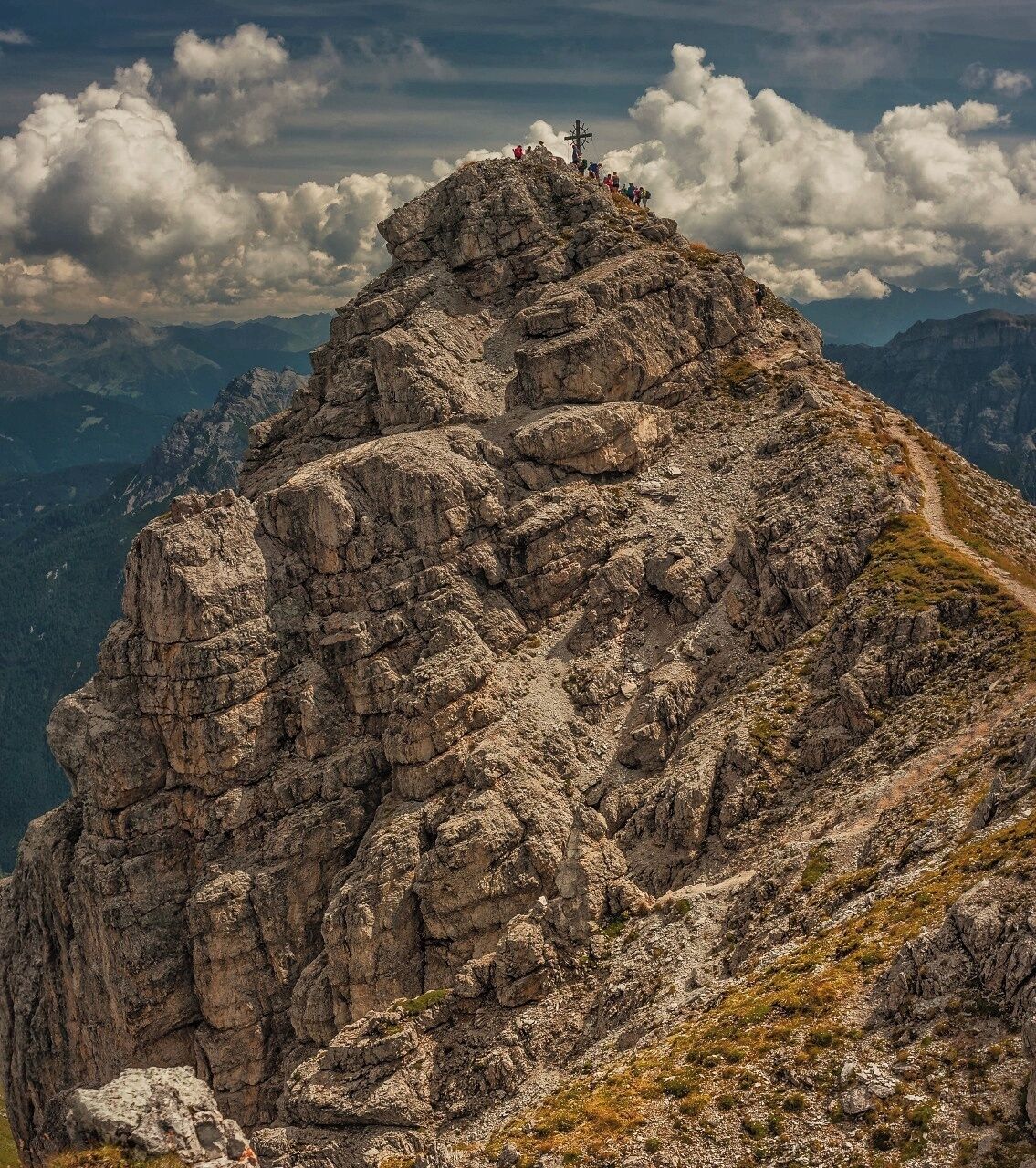 A rugged mountain peak with a cross at the summit and a trail leading up.