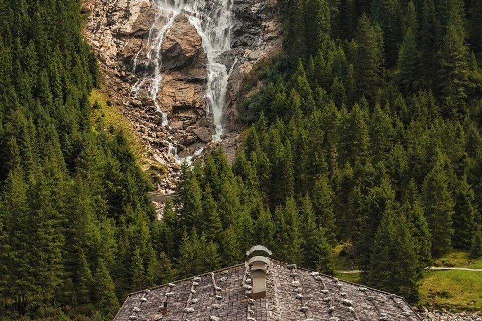 Een houten blokhut met een schuin dak, een waterval op de achtergrond en een bebost berggebied.