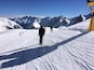 A snowy mountain slope with skiers, a clear blue sky, and distant snow-capped peaks.