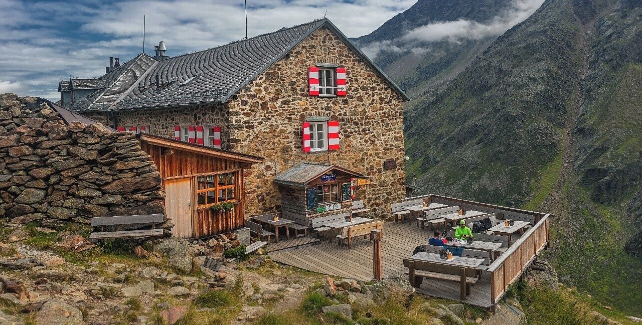 A stone building with red shutters, wooden deck, and mountainous backdrop.